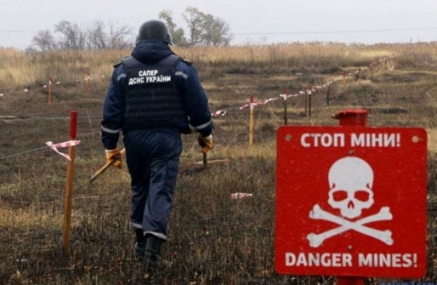 На відкритому полі спиною до глядача йде сапер у темно-синьому захисному спорядженні з написом «САПЕР ДСНС УКРАЇНИ» на спині, оглядаючи розмічену кілками та стрічками територію, а на передньому плані праворуч встановлено червону попереджувальну табличку з білим черепом і написами «СТОП МІНИ!» та «DANGER MINES!»