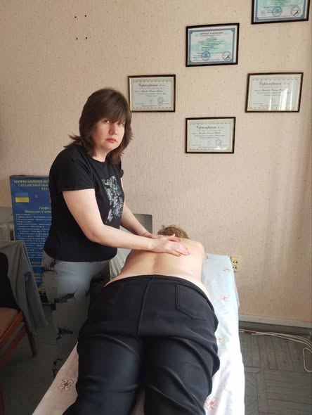 Жінка-масажист у чорній футболці зосереджено проводить сеанс масажу спини пацієнту на спеціальній кушетці в кабінеті, де на світлій стіні розвішані численні професійні дипломи та сертифікати у рамках