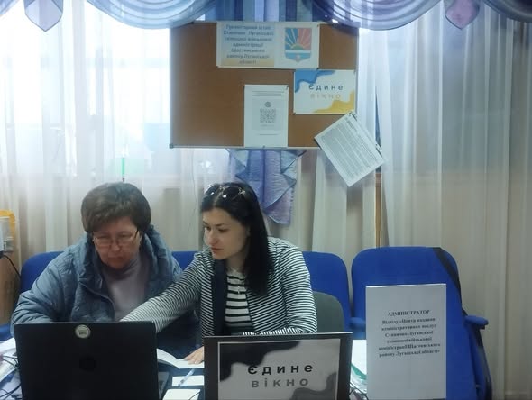 У світлій кімнаті з синіми кріслами дві жінки сидять за робочим столом перед ноутбуками біля таблички з написом «Єдине вікно», а позаду них на стіні висить дошка оголошень із гербом Луганської області.