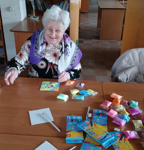 Літня жінка з білим волоссям і доброзичливою усмішкою, одягнена в чорну кофту з квітковим принтом і білу хустку з великими квітами та фіолетовим краєм, сидить за дерев'яним столом у приміщенні бібліотеки, де перед нею розкладено різноколірні пакетики з пластиліном або піском, олівці та невелика готова картинка з квітами