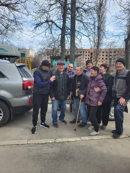 На асфальтованому майданчику біля сірого позашляховика стоїть група з восьми людей різного віку в темному осінньому одязі, які посміхаються та спілкуються між собою, а на задньому плані видно голі весняні дерева, дитячий майданчик та пошкоджену багатоповерхову будівлю.