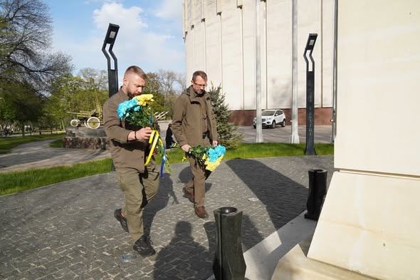 На тлі великої світло-сірої циліндричної будівлі діорами та вуличної експозиції з військовою технікою, двоє чоловіків у тактичному одязі оливкового кольору зосереджено йдуть брукованою доріжкою до білого меморіалу, тримаючи в руках великі букети жовтих і блакитних квітів, перев’язані стрічками в національних кольорах