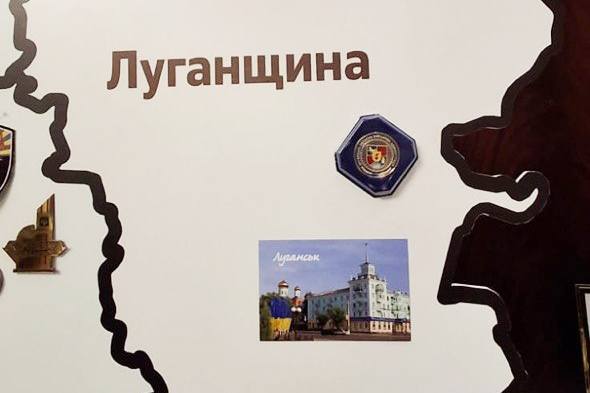 На світлій стіні розміщено частину експозиції з темним контуром карти області, де надруковано велике коричневе слово «Луганщина», а всередині закріплено синю шестикутну відзнаку з гербом та невелику горизонтальну фотографію з архітектурним пейзажем Луганська, що зображує світлу багатоповерхову будівлю зі шпилем та золоті куполи храму під ясним небом