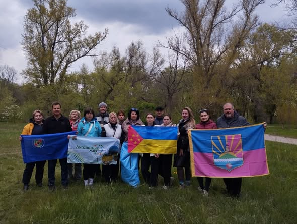 На груповій світлині, зробленій на тлі весняного лісу, зображено близько п'ятнадцяти людей, які позують на зеленій галявині, тримаючи перед собою державний прапор України та кілька прапорів територіальних громад чи організацій, зокрема великий малиново-синій прапор із гербом міста Лиман праворуч