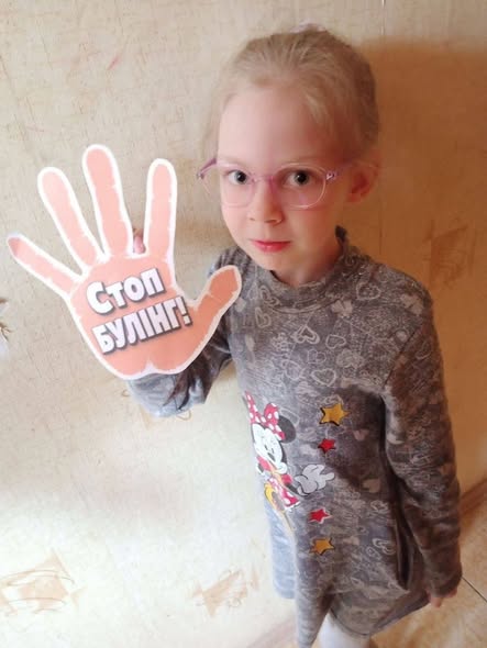 Маленька світловолоса дівчинка в рожевих окулярах і сірій сукні з Міккі Маусом стоїть біля світлої стіни, тримаючи в руці паперовий знак у формі долоні з написом «Стоп БУЛІНГ!», що символізує протест проти цькування.