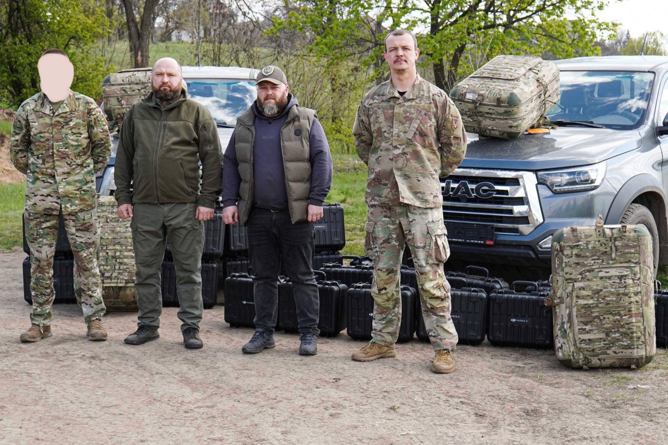 На фоні сірого пікапа та весняних дерев четверо чоловіків — двоє у військовому камуфляжі та двоє у тактичному цивільному одязі — стоять на ґрунтовій дорозі перед численними рядами чорних захисних кейсів і великих камуфляжних рюкзаків, при цьому обличчя крайнього зліва військовослужбовця приховано світлою плямою