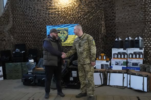 На фоні маскувальної сітки та прапора підрозділу «Помста» цивільний чоловік у жилеті та кепці тисне руку військовому в українському піксельному камуфляжі, стоячи серед численних коробок із портативними зарядними станціями Bluetti, акумуляторами та іншим технічним обладнанням.