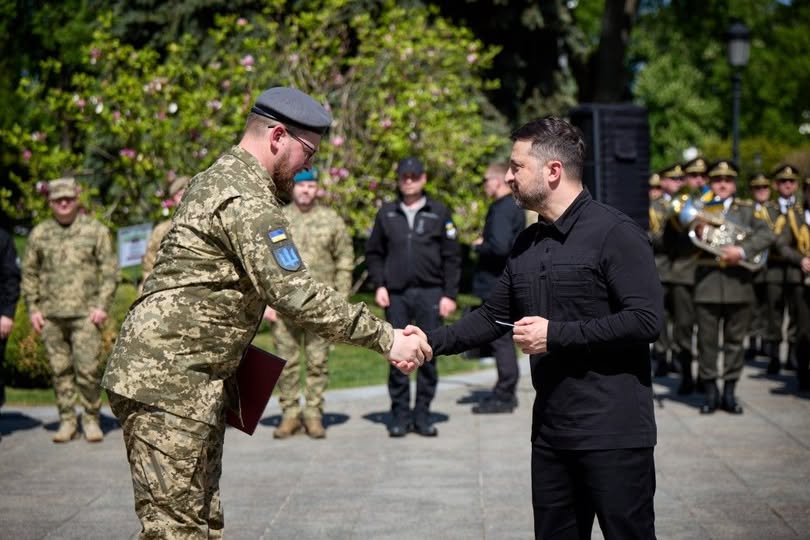 Президент України Володимир Зеленський тисне руку військовослужбовцю та вручає йому нагороду під час урочистої церемонії просто неба на фоні інших військових та дерев