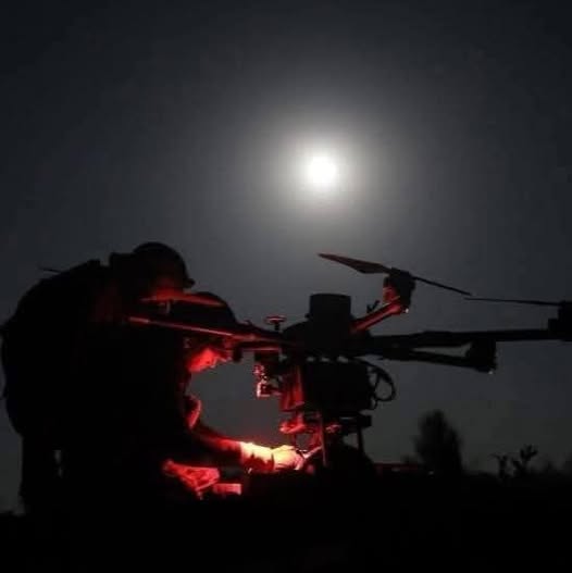 У нічній темряві під яскравим повним місяцем силуети двох операторів зосереджено готують до вильоту великий багатороторний дрон, деталі якого підсвічені контрастним червоним світлом для роботи в умовах маскування