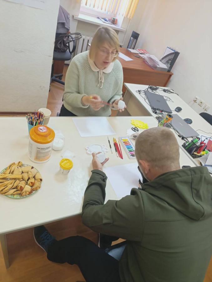 У світлому кабінеті за білим столом, заставленим ноутбуками, художнім приладдям та тарілкою з рогаликами, жінка в окулярах і світлому светрі натхненно демонструє чоловікові в оливковій куртці процес розпису білого яйця пензлем