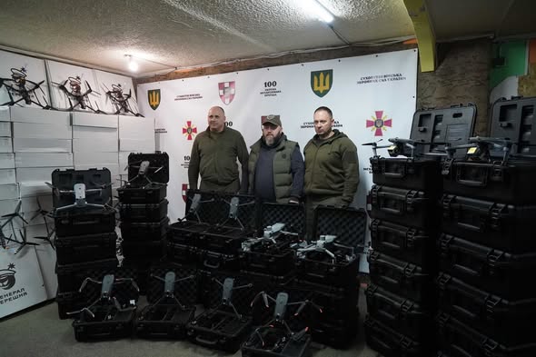 У приміщенні складу на фоні білого банера з офіційною символікою українських військових підрозділів троє чоловіків у тактичному одязі стоять в оточенні великої партії допомоги, що складається з численних квадрокоптерів у відкритих чорних кейсах та високих стопок білих коробок із технікою