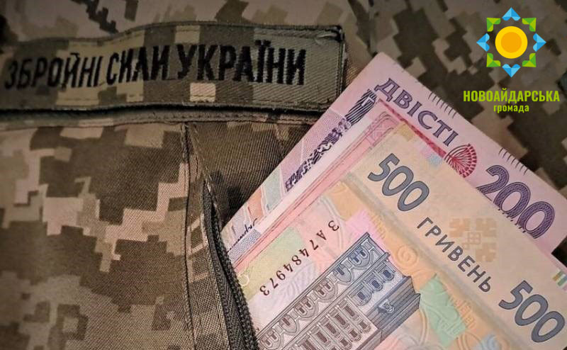 Кілька банкнот українських гривень номіналом двісті та п'ятсот стирчать із кишені камуфляжної військової форми з нашивкою «Збройні Сили України», а у верхньому правому куті розміщено логотип із написом «Новоайдарська громада»
