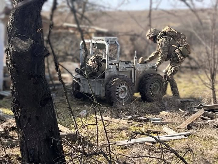 На сонячній відкритій місцевості серед сухої трави та залишків деревини солдат у камуфляжі та повному тактичному спорядженні нахиляється над невеликою чотириколісною роботизованою платформою, завантаженою речами, в той час як на лівому передньому плані височіє темний стовбур дерева