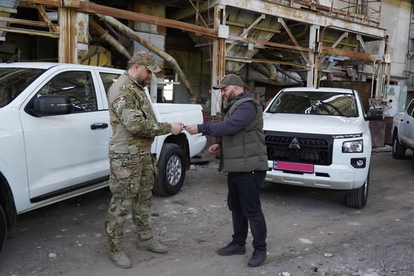 У промисловій зоні на фоні металевих конструкцій чоловік у темному жилеті та кепці передає ключі від нового білого пікапа Mitsubishi L200 військовослужбовцю у камуфляжі, а поруч стоїть ще один аналогічний автомобіль, що підкреслює момент передачі техніки для потреб оборони