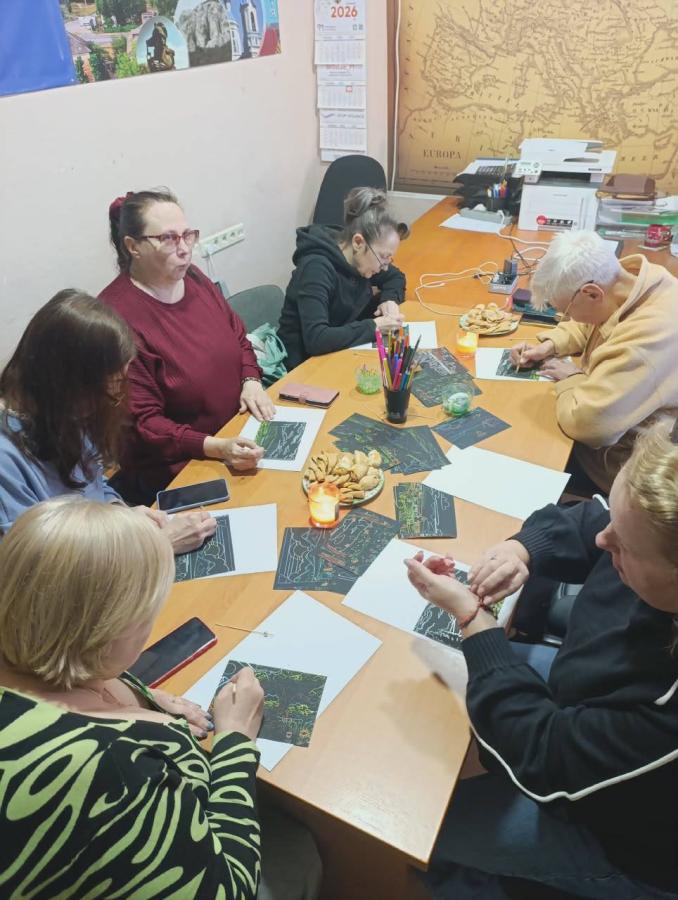 У невеликому кабінеті з мапою Європи та календарем на стіні п’ятеро жінок різного віку сидять навколо довгого дерев’яного столу, зосереджено працюючи над творчими малюнками в техніці подряпування на чорному фоні, а поруч із ними стоять стакан з олівцями, тарілка з печивом та запалена свічка