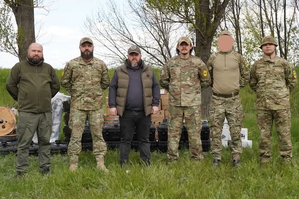 Шестеро чоловіків, четверо з яких одягнені в український камуфляж, а двоє — у тактичний цивільний одяг, стоять у ряд на зеленій галявині перед стопками чорних захисних кейсів і коробок із обладнанням на фоні високих дерев, причому обличчя одного з військовослужбовців розмите