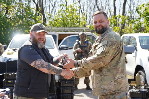 На сонячній галявині на фоні білих пікапів бородатий чоловік у цивільному жилеті та кепці з татуйованими руками передає ключі та тисне руку військовослужбовцю у камуфляжній формі, у той час як на задньому плані стоїть солдат зі зброєю, а поруч на землі складено чорні кейси з обладнанням