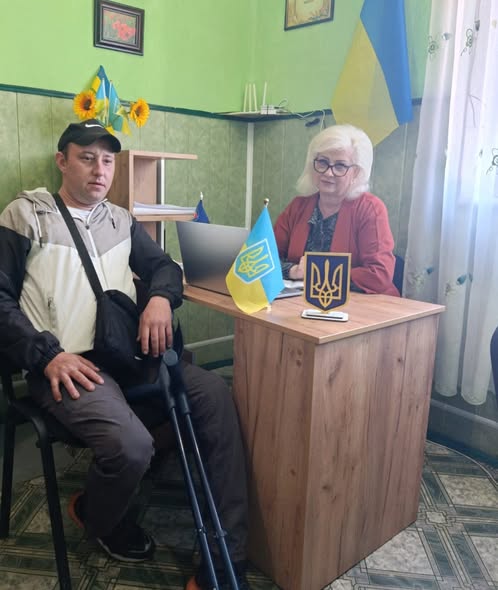 У кабінеті з зеленими стінами, декорованому державними символами України, за робочим столом із ноутбуком сидить жінка в червоному піджаку, а поруч на стільці — чоловік у спортивній кепці, який тримає металеву милицю.