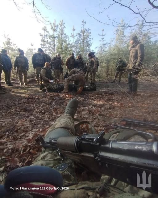 На знімку, зробленому від першої особи з позиції лежачи, видно руки військового з автоматом на фоні групи бійців у камуфляжі, які зібралися в осінньому лісі навколо пораненого побратима для надання допомоги