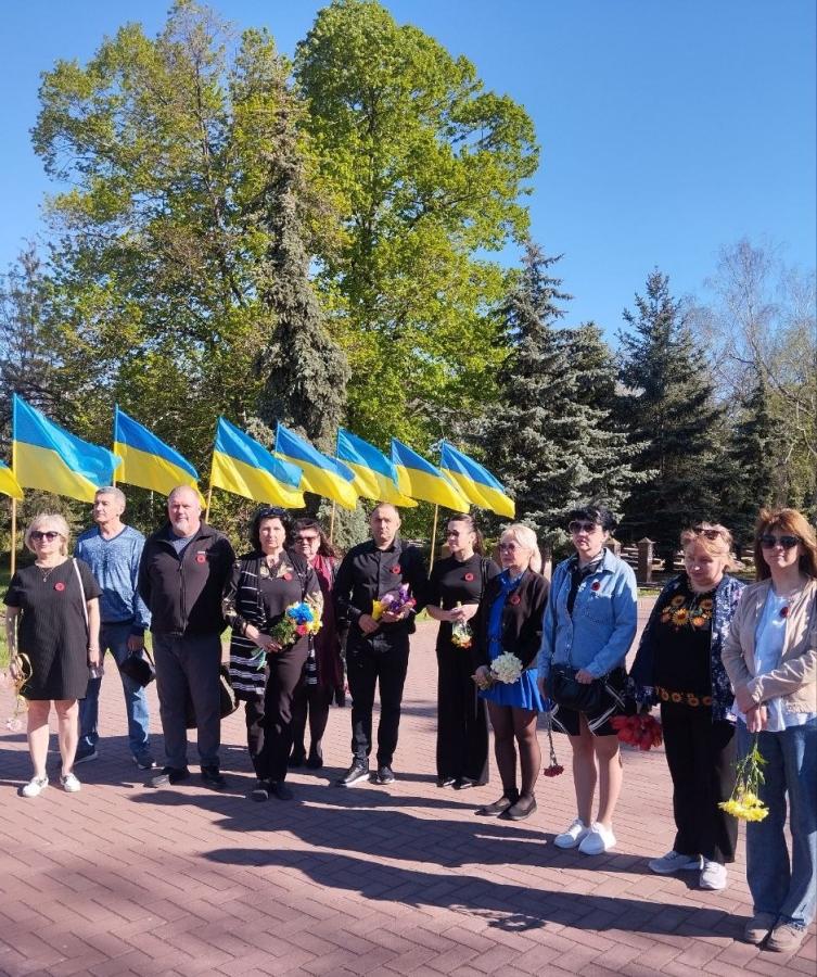 Близько одинадцяти людей різного віку стоять у шеренгу на брукованій доріжці. Майже у кожного на грудях прикріплена червона квітка маку — символ пам'яті. У руках деякі учасники тримають квіти (жовті та червоні гвоздики, синьо-жовті букети)