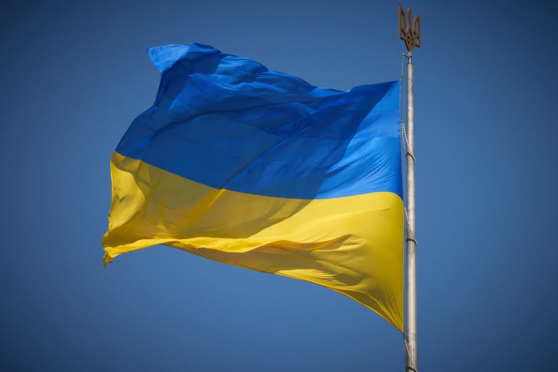 Державний прапор України з двох рівновеликих горизонтальних смуг синього і жовтого кольорів майорить на вітрі на високому металевому флагштоку, верхівку якого прикрашає золотистий тризуб на тлі чистого безхмарного неба