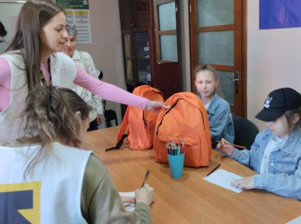Зліва стоять двоє дівчат-волонтерок. Одна з них, з довгим русявим волоссям, одягнена у світлий жилет поверх рожевої кофти. Вона простягає руку до яскраво-помаранчевого рюкзака, що стоїть на столі. Інша волонтерка, яку видно зі спини, також у білому жилеті з жовто-чорним логотипом, щось занотовує на папері