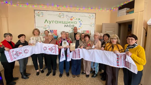 Люди стоять у два ряди. Перед собою вони тримають довгі білі рушники з червоно-чорною вишивкою, що тягнуться вздовж усієї групи. Деякі учасники тримають у руках невеликі книжки або брошури