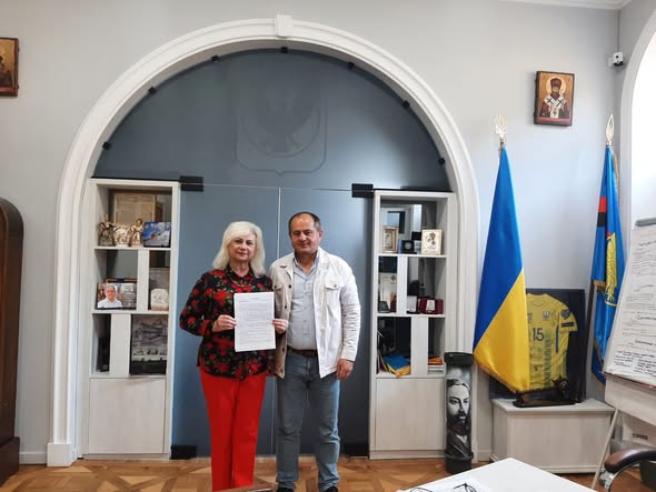 Жінка у яскравих червоних штанах і квітчастій блузі та чоловік у світлому піджаку стоять у кабінеті на фоні великої декоративної арки, полиць із пам'ятними речами та державного прапора України, разом тримаючи перед собою офіційний документ.
