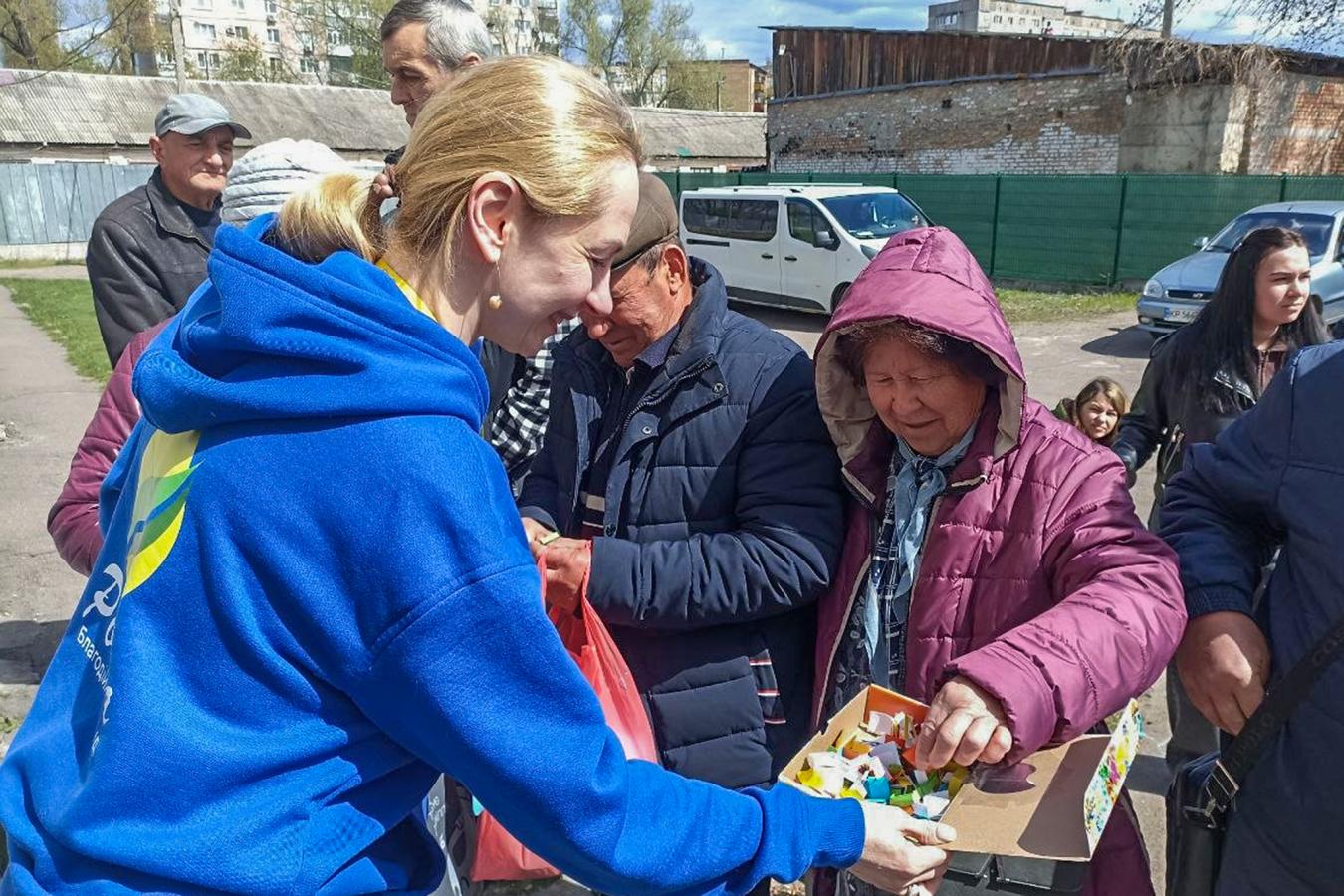 Молода жінка-волонтерка зі світлим волоссям, зібраним у хвіст, стоїть спиною до глядача ліворуч. Вона одягнена у яскраво-синю толстовку (худі) з жовто-зеленим логотипом на спині. Вона з посмішкою простягає відкриту картонну коробку з різнокольоровими дрібними предметами (схоже на цукерки в обгортках) людям перед нею