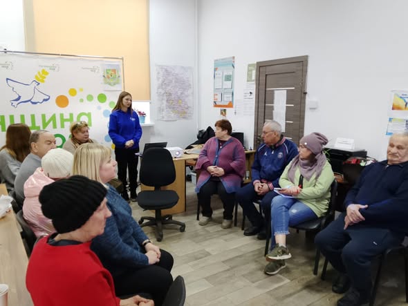 У світлому кабінеті група людей старшого віку сидить півколом і уважно слухає дівчину в синій кофті, яка стоїть біля ноутбука та проєктора на фоні інформаційного стенда з логотипом у вигляді голуба