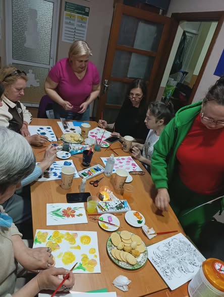 Група жінок різного віку зосереджено займається малюванням за довгим дерев'яним столом, на якому серед розкладених фарб, пензлів та склянок лежать готові малюнки з яскравими квітами, чисті аркуші та тарілки з частуванням