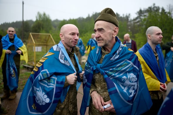 На передньому плані світлини під відкритим похмурим небом зображено двох усміхнених чоловіків у військовій формі, загорнутих у сині брендовані прапори з українською символікою та написами, причому ліворуч стоїть лисий військовий середнього віку, а праворуч — старший чоловік у зеленій пілотці з пачкою сигарет у руці, тоді як на задньому плані видно інших побратимів із синьо-жовтими прапорами на тлі зеленого лісу.