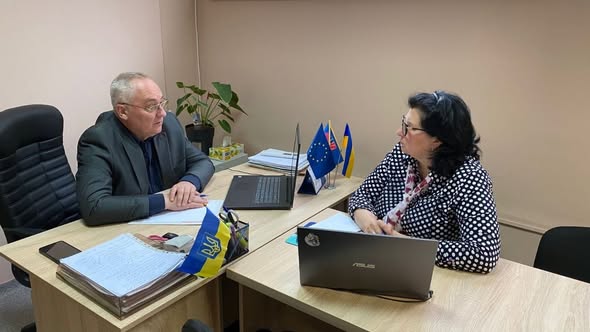 У робочому кабінеті за двома зсунутими дерев'яними столами, де стоять два відкриті ноутбуки, стоси документів та маленькі прапори України і Євросоюзу, сидить літній сивіючий чоловік у сірому піджаку та окулярах, який дивиться і розмовляє з чорнявою жінкою в окулярах та чорно-білій блузці в горошок, що сидить навпроти нього