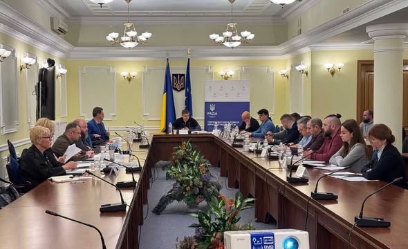 У світлій офіційній залі за великим П-подібним дерев'яним столом з настільними мікрофонами, документами та квітковими композиціями в центрі проходять збори або засідання комітету, де понад двадцять учасників у цивільному одязі сидять уздовж столів, а в торці зали під прапорами України та Європейського Союзу і гербом на стіні головує чоловік у темному одязі.