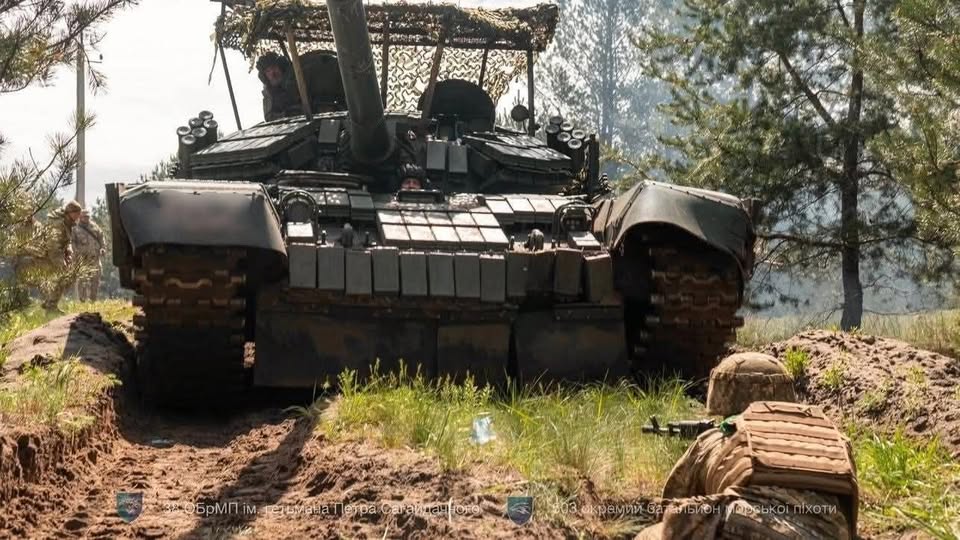На лісовій ґрунтовій дорозі великим планом спереду зображено український танк, оснащений динамічним захистом та антидроновою сіткою-навісом, з люків якого виглядають члени екіпажу, тоді як на передньому плані праворуч спиною до глядача в окопі лежить військовий у касці та бронежилеті зі зброєю, а ліворуч удалині стоять ще двоє бійців.