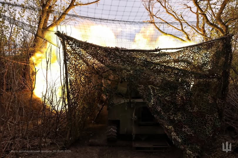 На світлині зафіксовано момент потужного спалаху вогню та диму від пострілу артилерійської установки, яка замаскована військовою сіткою серед дерев, із захисним сітчастим навісом зверху та офіційними написами у нижніх кутах