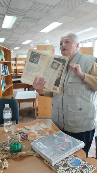 У приміщенні бібліотеки з дерев'яними книжковими стелажами та підвісною стелею сивочола жінка у світло-сірому жилеті з накладними кишенями стоїть біля столу і демонструє розгорнуту книгу з ілюстраціями круглих візерунків та текстом, у той час як на передньому плані на дерев'яній стільниці розкладено книги, прозору пластикову пляшку з водою, окуляри та мотки ниток і шнурів для рукоділля
