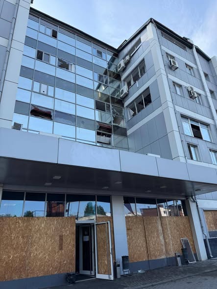 На світлині під кутом знизу вгору зображено багатоповерхову сучасну будівлю сірого кольору з великим скляним фасадом, на якому видно численні вибиті вікна та пошкодження, при цьому весь перший поверх та вхідна зона закриті захисними дерев'яними ОСБ-плитами, а на вищих поверхах помітні зовнішні блоки кондиціонерів