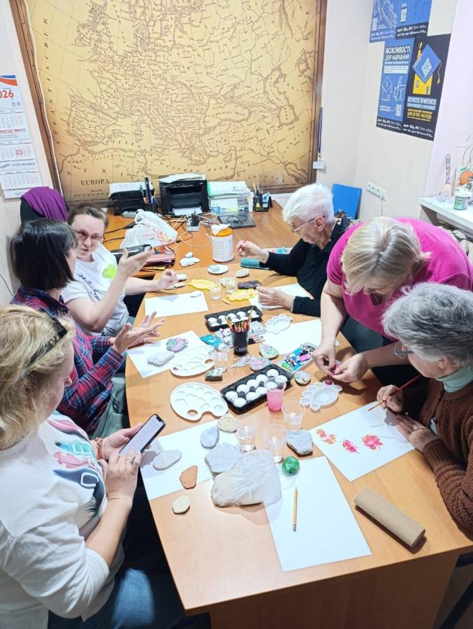 На світлині згори зафіксовано творче заняття, під час якого семеро жінок різного віку сидять за довгим дерев'яним столом у кабінеті на тлі великої старовинної карти Європи і розписують фарбами камінці та папір, використовуючи художні пензлі, пластикові палітри, стаканчики з водою та олівці.