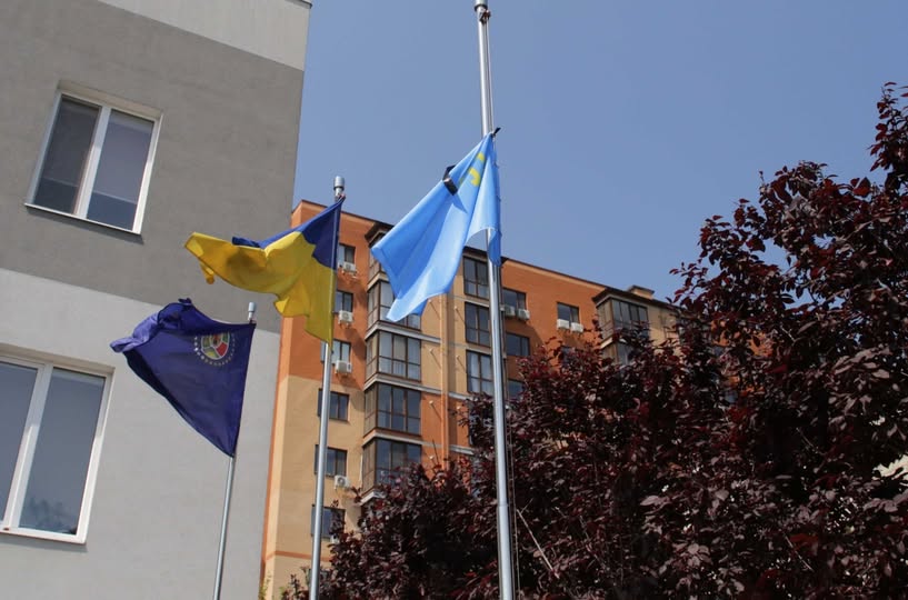 На світлині знизу вгору на тлі чистого синього неба зображено три прапори на високих металевих флагштоках — темно-синій прапор із круглою емблемою ліворуч, державний синьо-жовтий прапор України по центру та блакитний прапор із золотою кримськотатарською тамгою праворуч, при цьому на знак жалоби до верхньої частини кримськотатарського стяга прикріплено чорну стрічку, а на задньому плані видно частину сучасної житлової будівлі та густу крону дерева з темно-бордовим листям.