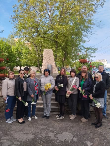 На світлині зображено групу з одинадцяти людей, які стоять півколом на вулиці біля кам'яного пам'ятного знака в оточенні зелених дерев, при цьому майже всі присутні тримають у руках весняні квіти, такі як нарциси, тюльпани та гвоздики