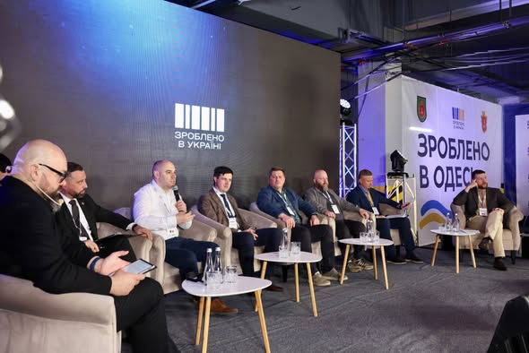 Група чоловіків у костюмах сидить у кріслах на сцені під час офіційного заходу на тлі великих екранів із написами «Зроблено в Україні» та «Зроблено в Одесі»