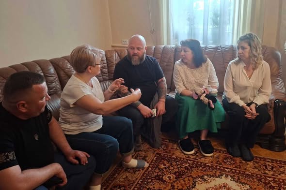 Група з п'яти людей сидить на великому коричневому шкіряному дивані та поруч із ним у кімнаті з килимом на підлозі, активно спілкуючись між собою, при цьому жінка у білій футболці на передньому плані жестикулює руками
