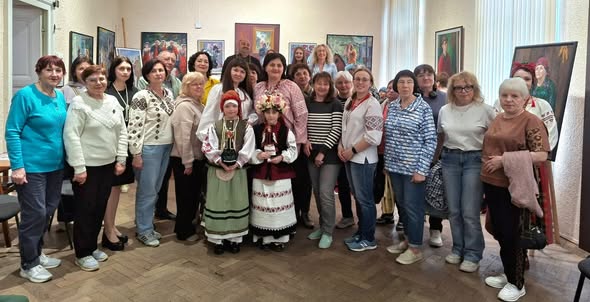 Велика група людей, переважно жінок у сучасному та традиційному українському одязі, стоїть разом у музейній залі на тлі розвішаних на стінах картин, а в центрі першого ряду знаходяться двоє дівчаток у вишиванках та вінках з обрядовими ляльками-мотанками в руках
