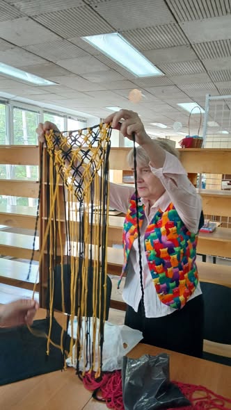 Жінка у яскравому різнокольоровому жилеті стоїть біля дерев'яного стелажа і піднімає руками плетений виріб із довгими жовтими та чорними нитками макраме