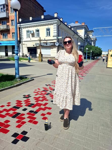 Молода жінка у світлій сукні в дрібний квіточку, сонцезахисних окулярах та чорних рукавичках стоїть посеред пішохідної вулиці міста поруч із намальованим на бруківці великим червоно-чорним орнаментом вишиванки, тримаючи в руках паперовий стаканчик.