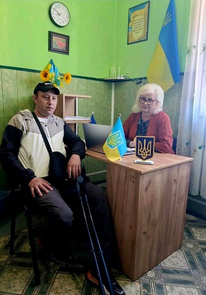 У кабінеті із зеленими стінами чоловік у кепці та з милицями сидить на стільці поруч із дерев'яним робочим столом, за яким сидітиме жінка в окулярах та червоному піджаку на тлі державного прапора України.
