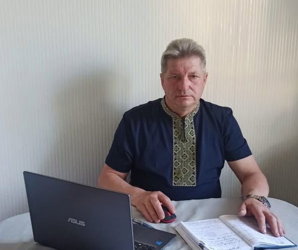 Чоловік середнього віку із сивим волоссям у темно-синій сорочці з вишитим коміром та планкою сидить за столом перед відкритим ноутбуком ASUS, тримаючи руку на комп'ютерній миші поруч із розгорнутим блокнотом