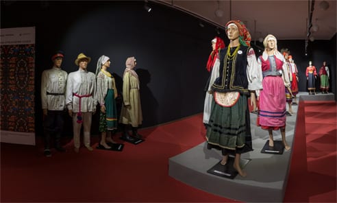 На зображенні представлено музейну експозицію з манекенами в традиційному українському народному вбранні різних регіонів, що розміщені у виставковій залі з темними стінами та червоною підлогою