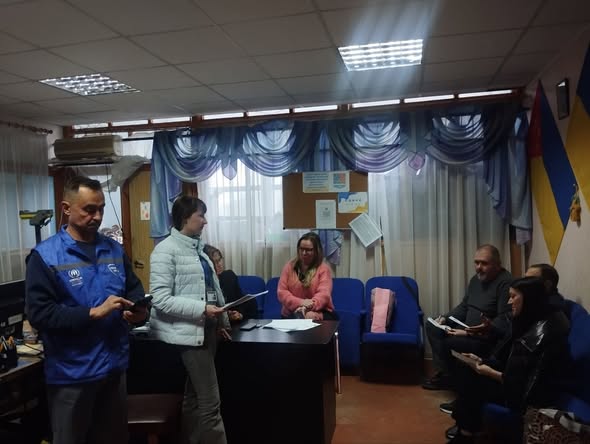 У кабінеті з великим вікном, завішеним блакитним тюлем, та українським прапором на стіні зафіксовано робочу зустріч кількох людей, де частина присутніх сидить у синіх кріслах, а жінка у світлій куртці та чоловік у синьому жилеті УВКБ ООН стоять біля письмового столу